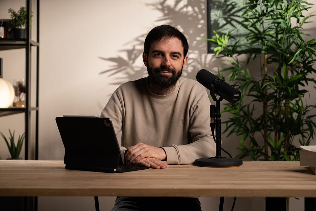 Homme barbu en sweat beige assis à un bureau, face caméra, prêt à enregistrer avec un micro podcast.