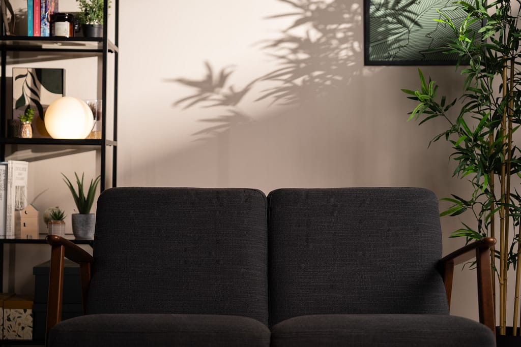 Détail du décor L’Essentiel avec canapé gris en avant-plan et ombres nettes de feuilles projetées sur le mur, créant une ambiance chaleureuse.