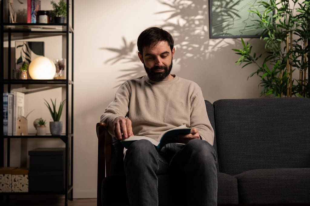Personne lisant un livre, assise sur un canapé gris dans le décor L’Essentiel, avec une lumière douce projetant l’ombre d’un feuillage sur le mur.