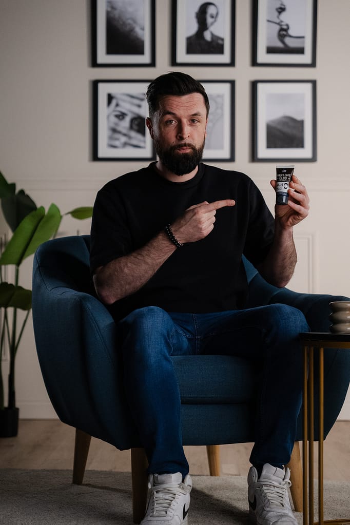 Homme assis dans un fauteuil bleu qui pointe du doigt un tube de crème dans le décor La Parenthèse.