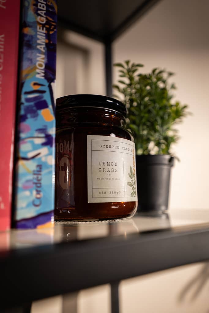 Gros plan sur une bougie parfumée “Lemongrass” posée sur une étagère, entre des livres et une plante verte.