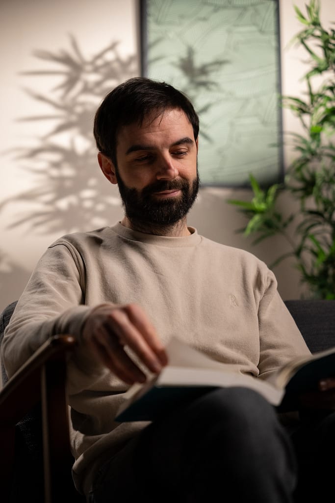 Homme feuilletant un livre, assis dans un fauteuil, lumière douce et ambiance végétale.
