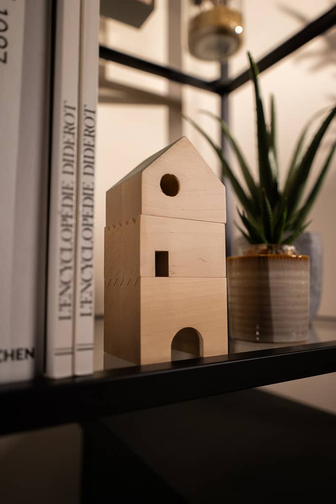 Maison miniature en bois posée sur une étagère, entourée de livres et de plantes déco.